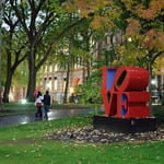 Photo of paved roadway leading from lower left to center, with brick university building in background, and red LOVE sculpture in the foreground.